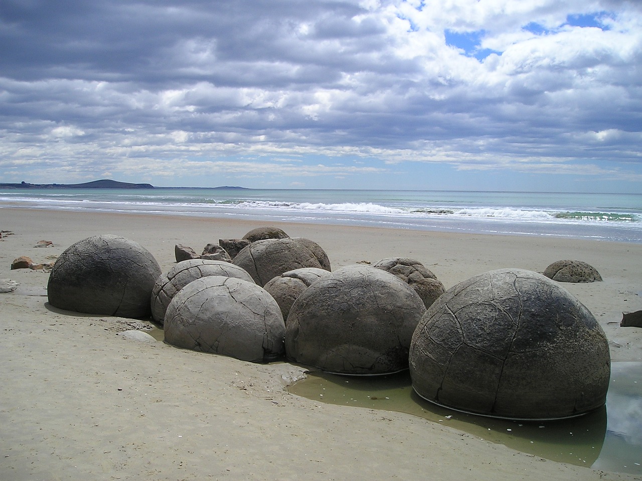 Gerundete Steine an einem Sandstrand, im Hintergrund das Meer und ein bewölkter Himmel